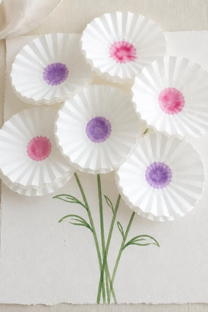 A flat-lay photo of a bouquet made from multiple white coffee filters layered like petals. Each filter has a small watercolor dot center in pink and purple, and green stems are drawn with marker.