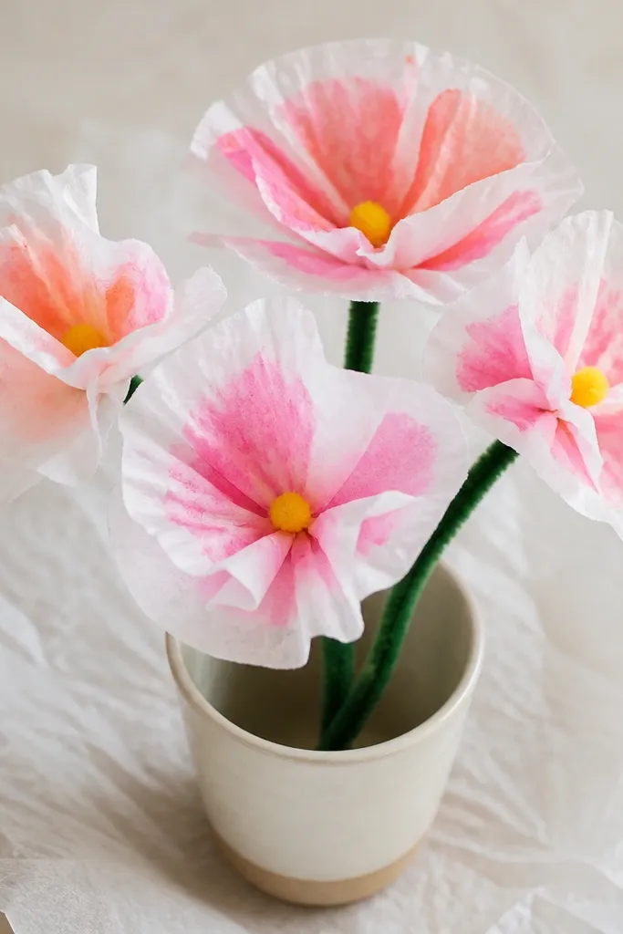 Several white coffee filter flowers on a sheet of wax paper; each bloom has a watercolor wash in pink and coral on the petals with tiny yellow centers; a green pipe cleaner stem holds them in a little cup.