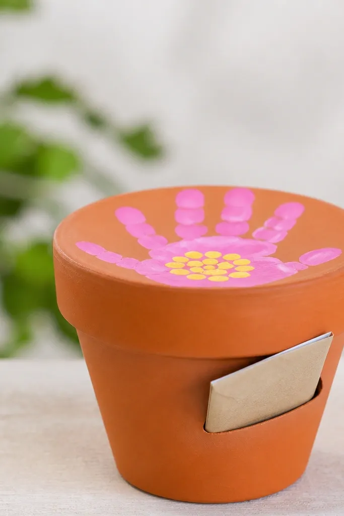 A small flower pot painted terracotta with a handprint flower on top; the handprints are bright pink with yellow dots; a mini seed envelope is tucked into a slit on the side of the pot.