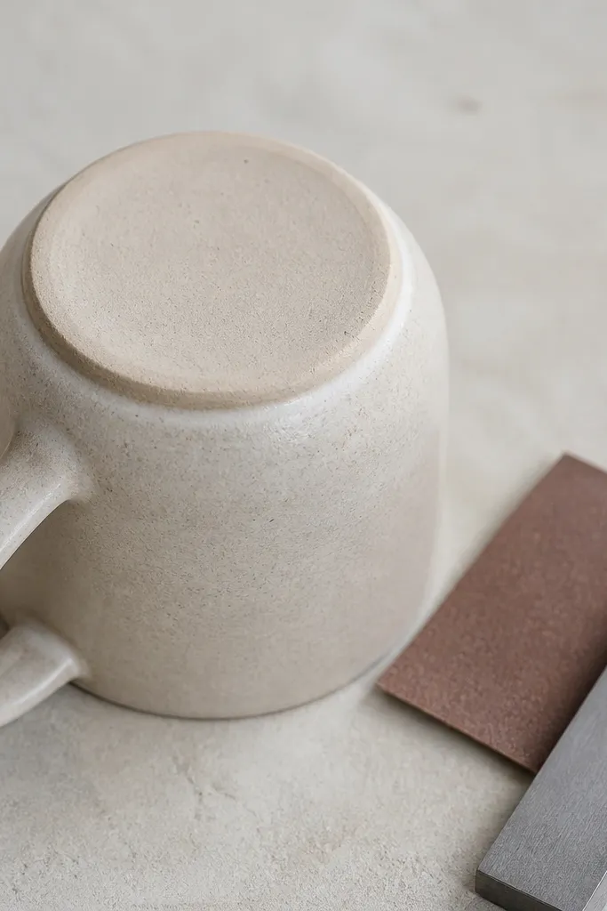 A mug turned upside down showing a neat foot ring; the bottom has been trimmed flat with a loop tool. A small piece of sandpaper and a straightedge are placed next to it.