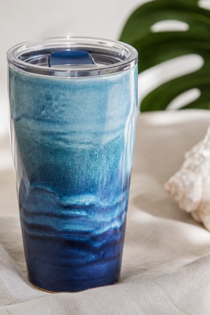 A handmade ceramic travel mug glazed in layered ocean blues with visible wave-like texture. The rim has a subtle ripple profile, and the lid is a clear plastic top with a blue base.