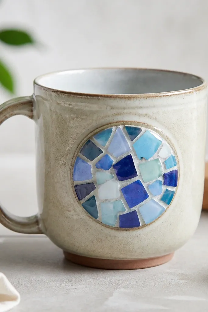 A mug with a small mosaic tile inlay on one side, each tile piece different shades of blue and white; the mosaic is covered with a smooth clear epoxy dome.