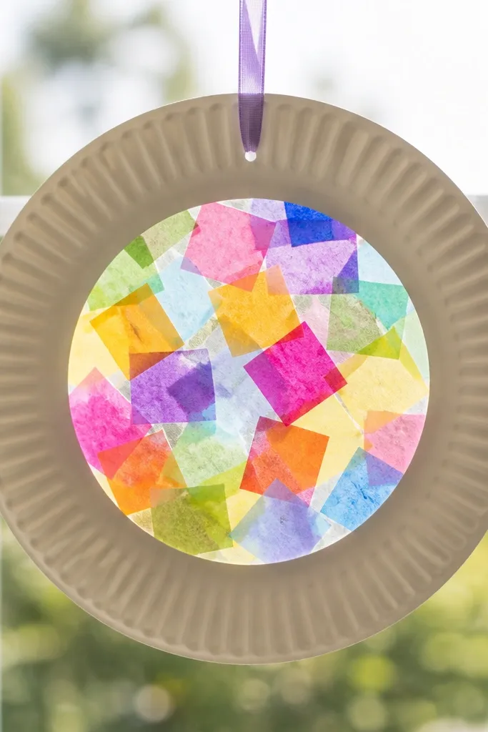 A paper plate sun-catcher with a hole in the center, tissue paper squares filling the middle, and a ribbon loop at the top, shown in front of a window for light glow.