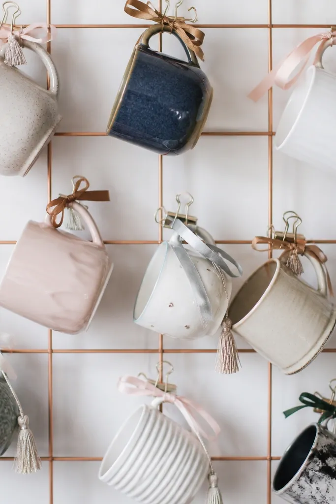 A cluster of mismatched mugs attached to a wire grid using binder clips, with tassels and ribbon tied around the mug handles.